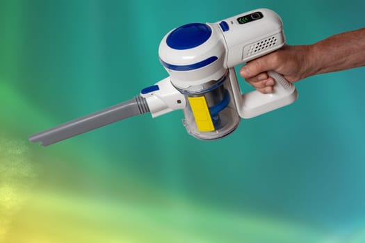The male hand holds hold a modern cordless battery-powered vacuum cleaner over abstract blurred background. Space. Household cleanliness and hygiene.