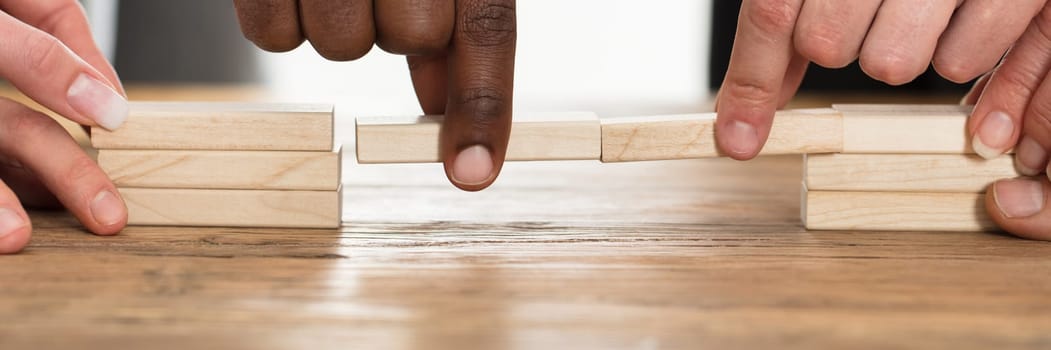 Businesspeople's Hand Holding Wooden Building Blocks To Form A Bridge Over A Gap On Table