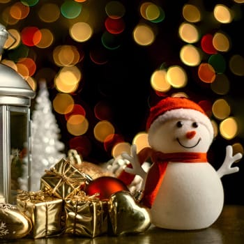 Christmas composition: A snowman, a lantern with candles, bright shiny boxes with gifts, blurred bokeh lights in the background.
