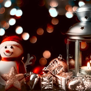 Christmas composition: A snowman, a lantern with candles, bright shiny boxes with gifts, blurred bokeh lights in the background.