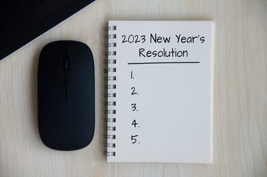 2023 New Year's Resolutions text on note pad with mouse and laptop on wooden desk.t