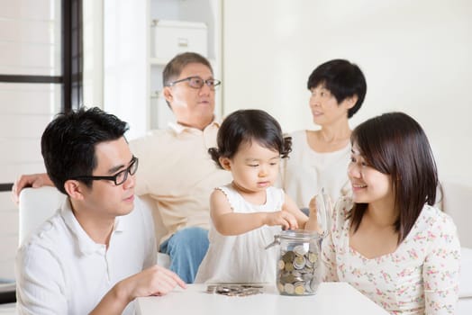 Toddler putting coins into money jar. Asian family money savings concept. Multi generations living lifestyle at home.
