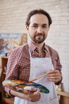 Modern artist with paintbrush and palette looking at camera in studio
