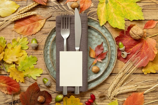 Autumn rustic table place with blank card between colorful leaves and berries, top view, mockup. Flat lay with vertical card on wooden table, copy space. Thanksgiving or harvest concept