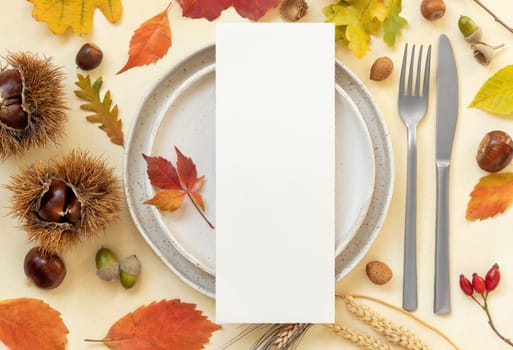 Autumn table place with colorful leaves and blank menu card top view. Mockup of fall vertical card on light yellowtable, flat lay, copy space. Harvest, Halloween and Thanksgiving season