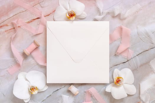 Wedding square envelope near white orchid flowers and silk ribbons on marble top view, mockup. Romantic scene flat lay. Valentines or Spring romantic concept
