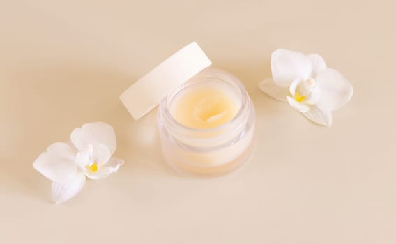 Small opened glass jar near white orchid flowers on light yellow close up, mockup. Skincare beauty product package. Exotic natural cosmetics, pastel minimal composition..