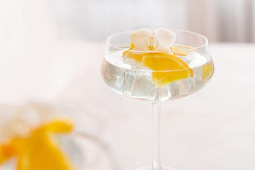 Transparent cocktail with ice cubes in a champagne coupe glass decorated with slices of orange white orchid flower close up. Tropical drink at the bar. Summer vacation mood