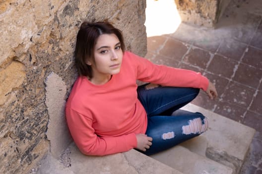 Teen girl wearing pink long sleeve shirt looking into a camera. Mockup. Serious Teenager girl sitting outdoor on a stairs spending her time alone. Adolescence concept