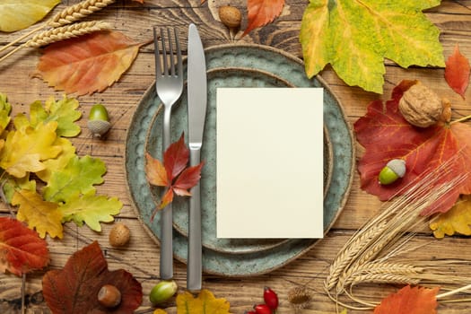 Autumn rustic table setting with blank card between colorful leaves and berries, top view. Vertical card mockup on wooden table, flat lay, copy space. Thanksgiving, halloween or harvest concept