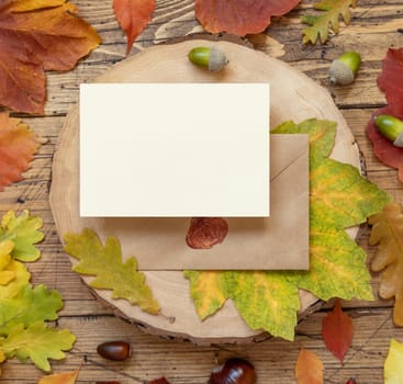 Blank card between colorful autumn leaves and berries on wooden table top view, rustic mockup. Horizontal card over envelope, flat lay, copy space. Thanksgiving, halloween or harvest concept