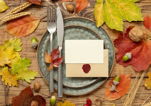 Autumn rustic table setting with blank place card and envelope between colorful leaves and berries, top view, mockup. Flat lay Horizontal card on wooden table, flat lay, copy space. Thanksgiving or harvest