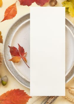 Autumn table place with colorful leaves and blank menu card top view. Mockup of fall vertical card on light yellowtable, flat lay, copy space. Harvest, Halloween and Thanksgiving season