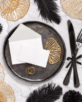 Black and golden wedding table setting with a place card and rings top view, mockup. Elegant flat lay with paper card and envelope, wedding or holiday stationery template