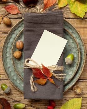 Autumn rustic table place with blank card between colorful leaves and berries, top view, mockup. Flat lay with vertical card on wooden table, copy space. Thanksgiving or harvest concept