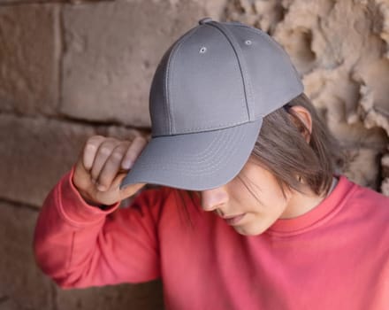 Teen girl in grey baseball cap and pink long sleeve shirt looking into a camera. Cap mockup. Serious Teenager girl near a stone wall outdoor touching the visor. Loneliness concept
