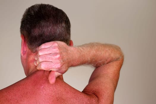 Mature adult white male seen from rear view massaging the back of his neck under his brown hair. Rear view of a naked caucasian man massaging his painful neck to help to relax his muscles close-up
