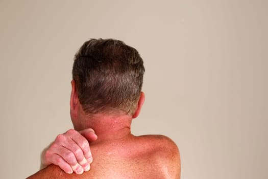 Right hand of a shirtless adult white man massaging his left shoulder as seen from a rear view. Close-up of a caucasian guy rubbing his left shoulder with his right hand in front of a wall indoors