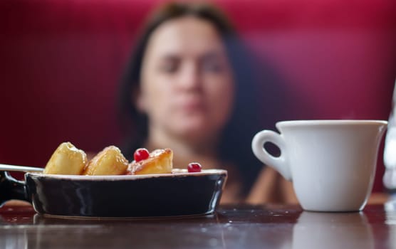 Portion of round paneer cheesecakes on a plate on a table with a white cup of coffee on a blurred background of an unrecognizable girl and restaurant interior. Healthy morning breakfast