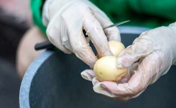 Woman's hands in white gloves peel raw new potatoes, close-up. Cleaning organic potatoes. Detail of female hands peeling fresh yellow potato with kitchen knife, food preparation concept