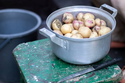 Stainless steel saucepan with raw new potatoes and a kitchen knife. Potatoes not peeled. Close-up of yellow and red skinned potatoes. Peeling potatoes in the kitchen on a wooden rustic table