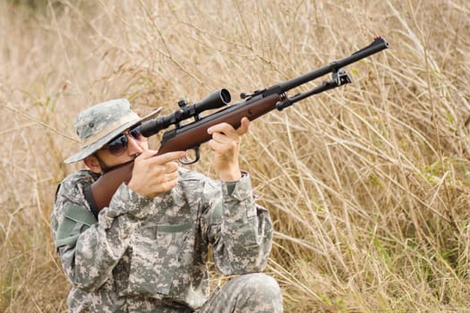 The hiding hunter in military uniform is aiming with shotgun or air gun outdoors in nature.