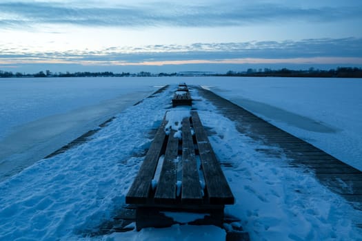 Wooden bench on a snow-covered pier and a frozen lake after sunset, Zoltance, Poland