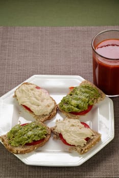 Four half slices of open faced sandwiches of hummus and tomato and guacamole and tomato on bread near a tomato vegetable beverage. Hummus, guacamole and tomato slices of four half slices of bread meal