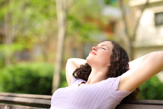 Relaxed woman resting with arms on head sitting in a bench in a park