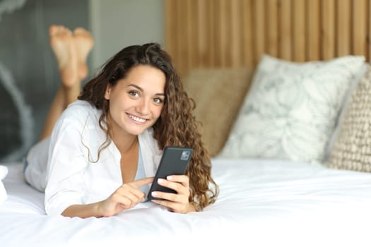 Happy woman holding smart phone looks at you lying on a bed