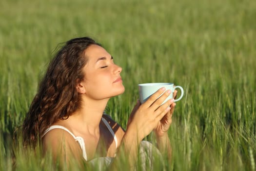 Woman relaxing smelling coffee from cup in a green wheat field