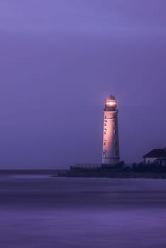 A lighthouse on the coast of the sea at dusk, its bright lamp is a landmark for ships. A lighthouse in the lilac twilight. Seascape, nature background.