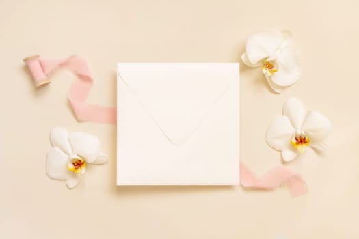 Wedding square envelope near white orchid flowers and silk ribbons on light yellow top view, mockup. Romantic scene with blank card flat lay. Valentines or Spring romantic concept