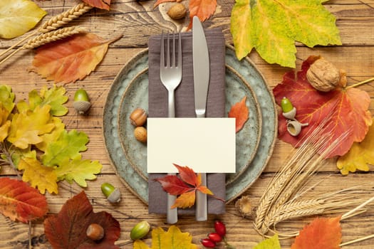 Autumn rustic table place with blank card between colorful leaves and berries, top view, mockup. Flat lay with horizontal card on wooden table, copy space. Thanksgiving or harvest concept