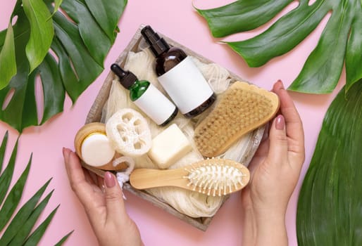Hands holding tray of Homemade Cosmetics and eco friendly accessories near tropical leaves, pink background,. Brand package mockup. Natural beauty products for hair and skin care, Top view