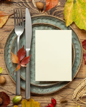 Autumn rustic table setting with blank card between colorful leaves and berries, top view. Vertical card mockup on wooden table, flat lay, copy space. Thanksgiving, halloween or harvest concept