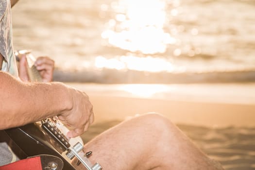 Romantic young man sitting on the sand at sunset with playing the guitar