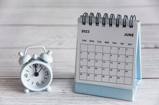 June 2023 white desk calendar on wooden table with alarm clock pointing at 12 o'clock.