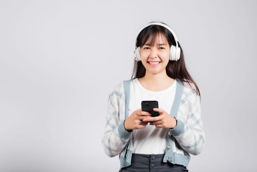 Woman excited smiling listening to music radio in bluetooth headphones and holding phone studio shot isolated white background, happy Asian young female listen modern technology mobile phone