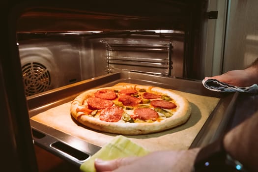 A man's hands pull out a homemade pepperoni pizza from the oven.