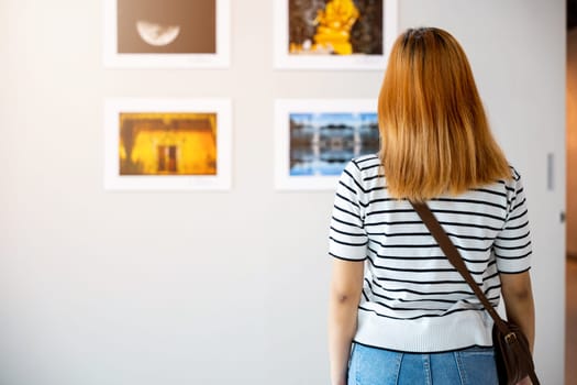 Asian woman standing she looking art gallery in front of colorful framed paintings pictures on white wall, young female watch at photo frame to leaning against at show exhibition gallery, Back view