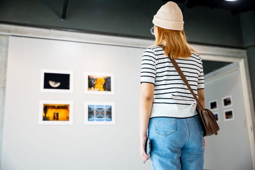 Asian woman standing she looking art gallery in front of colorful framed paintings pictures on white wall, people watch at photo frame to leaning against at show exhibition artwork gallery, Back view