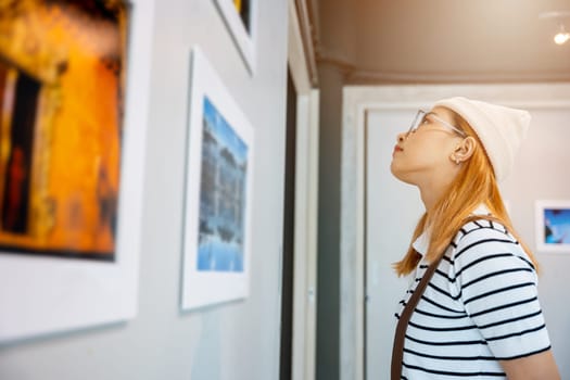 Asian woman standing she looking art gallery in front of colorful framed paintings pictures on white wall, people watch at photo frame to leaning against at show exhibition artwork gallery, Side view