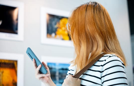 Visitor woman standing takes picture art gallery collection in front framed paintings pictures on white wall with mobile phone, people watch at photo frame with smartphone at artwork gallery show