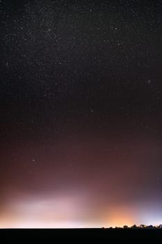 Night Starry Sky With Glowing Stars Above Countryside Town Landscape. Rural Field Meadow In Early Spring. Bright Glow Of Stars. Illumination Lights Of Small Town Or Village.
