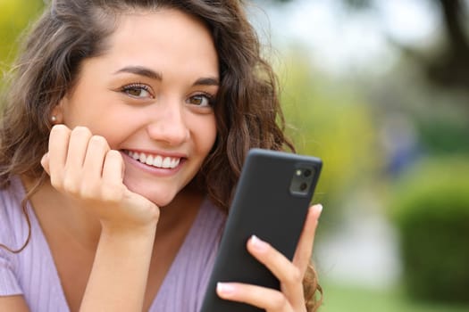 Happy beautiful woman holding cell phone looks at you in a park