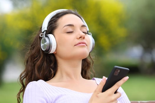 Relaxed woman wearing headphones and holding smart phone meditating listening guide in a park