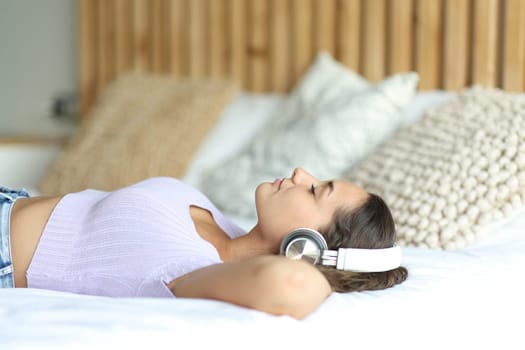 Side view portrait of a woman wearing wireless headphones relaxing listening to music on a bed in a room