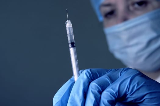 A woman doctor in a medical cap and mask holds a medical syringe in her hand. Soft focus..