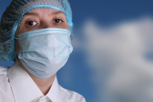 Close-up portrait of a woman doctor in a medical cap and mask against the blue sky..